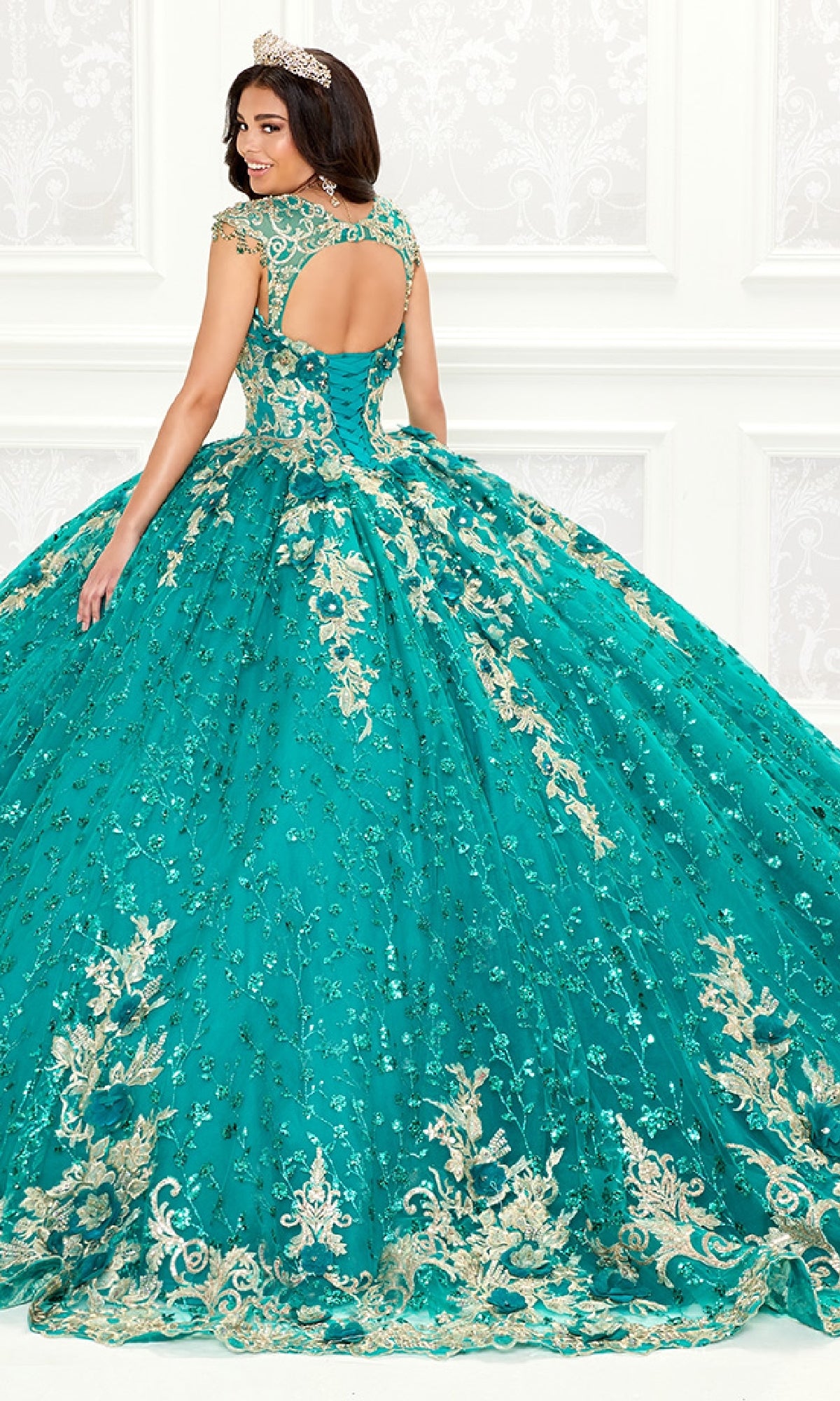 A woman in a jeweled tiara wears the Quinceanera Dress PR30088 By Princesa, featuring gold embroidery, lace details, and a full skirt, as she stands with her back to the camera in an ornate white room.